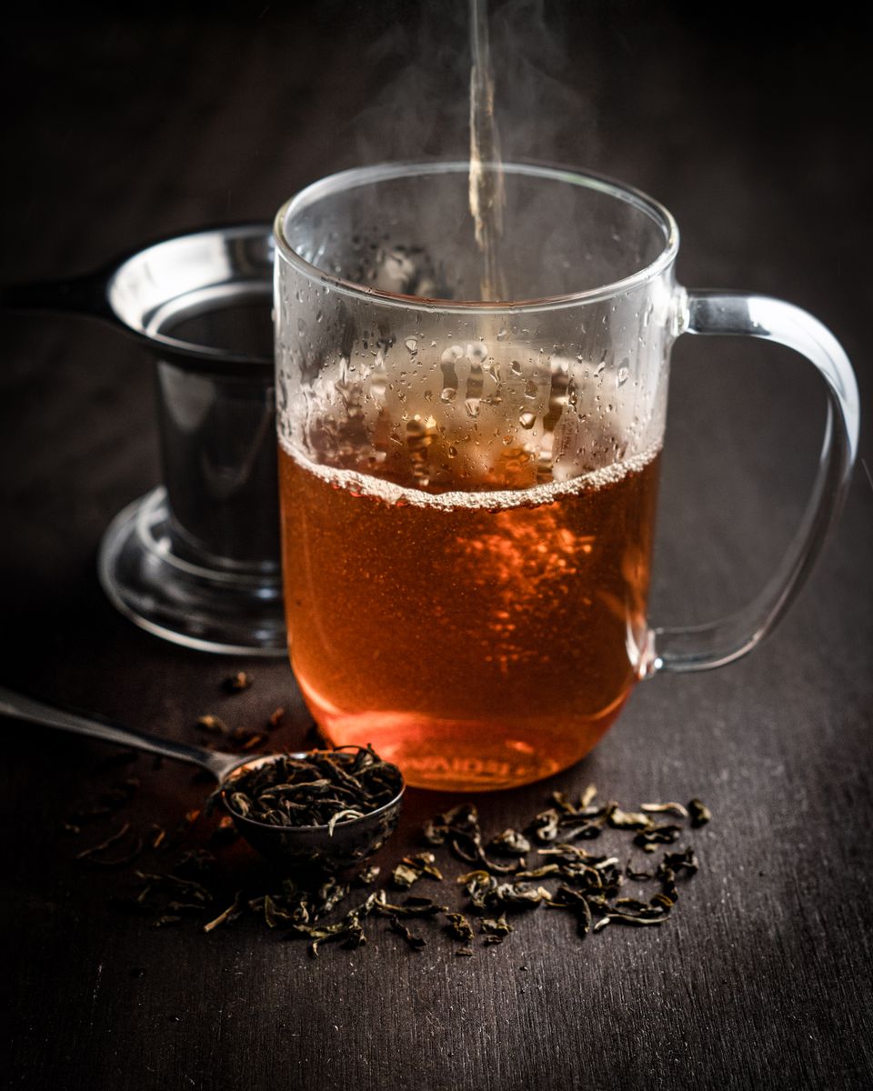 Illustrative photo of hot amber tea being poured into a clear glass mug beside loose tea leaves.