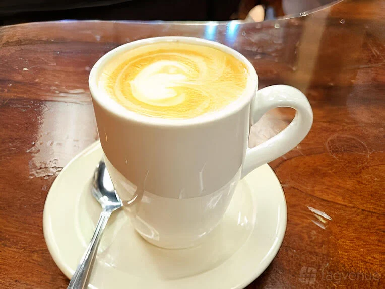 A milk coffee in a white mug and saucer on a wooden table at Zed’s Cafe.